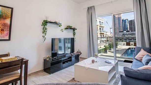 Un salon moderne doté d’un téléviseur à écran plat, d’un coin repas avec une table et des chaises en bois, d’une table basse blanche et d’une grande fenêtre offrant une vue sur la ville.
