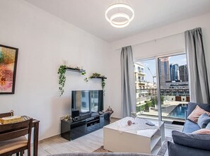 A modern living room with a flat-screen TV, a dining area with a wooden table and chairs, a white coffee table, and a large window offering a city view. - Sophisticated 1BR Apartment in Oxford Residence (Dubai)