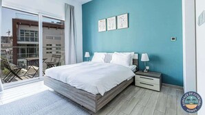 Une chambre moderne avec un grand lit, une table de chevet et une vue sur un balcon aménagé avec du mobilier d’extérieur.