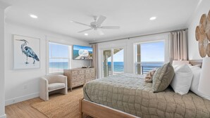 Een slaapkamer met een bed, een kaptafel, een stoel, een plafondventilator en een groot raam met uitzicht op de oceaan.