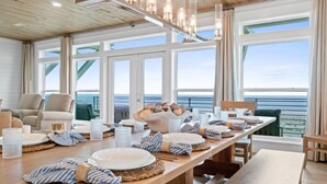 Een eetgedeelte met een lange houten tafel gedekt voor een maaltijd, grote ramen met uitzicht op zee, en een gezellige zithoek met een bank en stoelen.