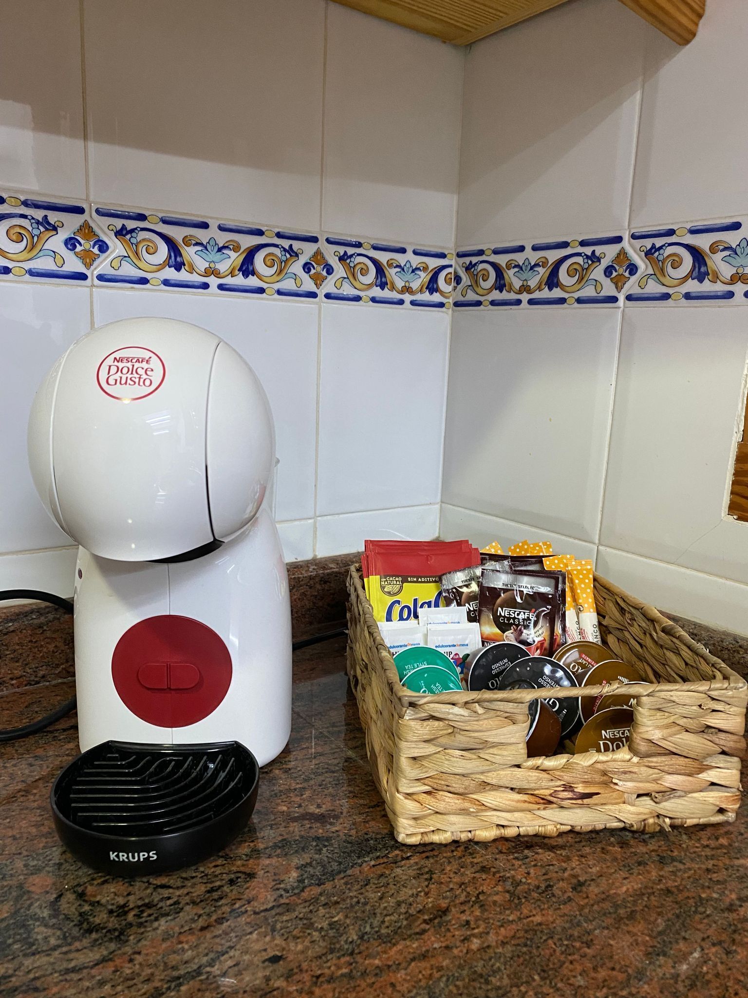 Ett kök med en vit köksmaskin, en korg med Nescafé Dolce Gusto-kapslar och en Krups-kaffebryggare.