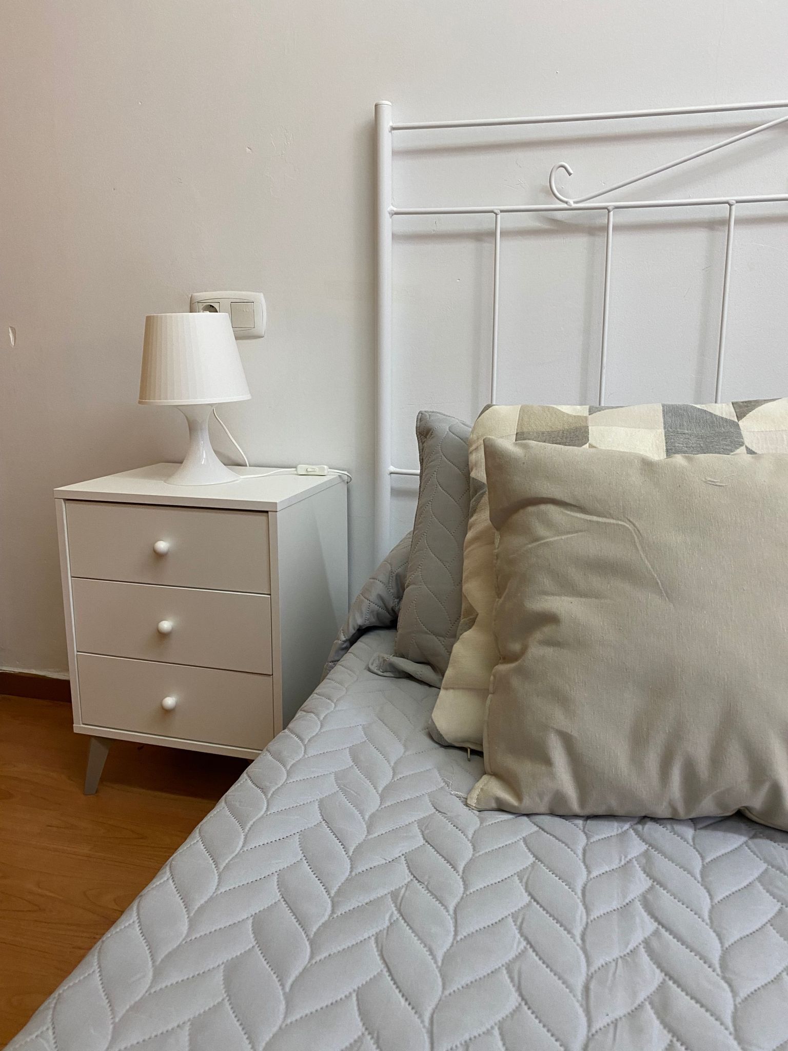 Un dormitorio ordenado con cama, mesita de noche con lámpara y cabecero fijado a la pared.