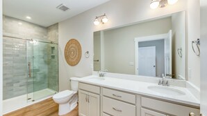 A modern bathroom with a glass shower enclosure, a toilet, a large mirror, and a vanity with a sink. - Hamilton Cay Reserve at Bermuda Bay (Kill Devil Hills)