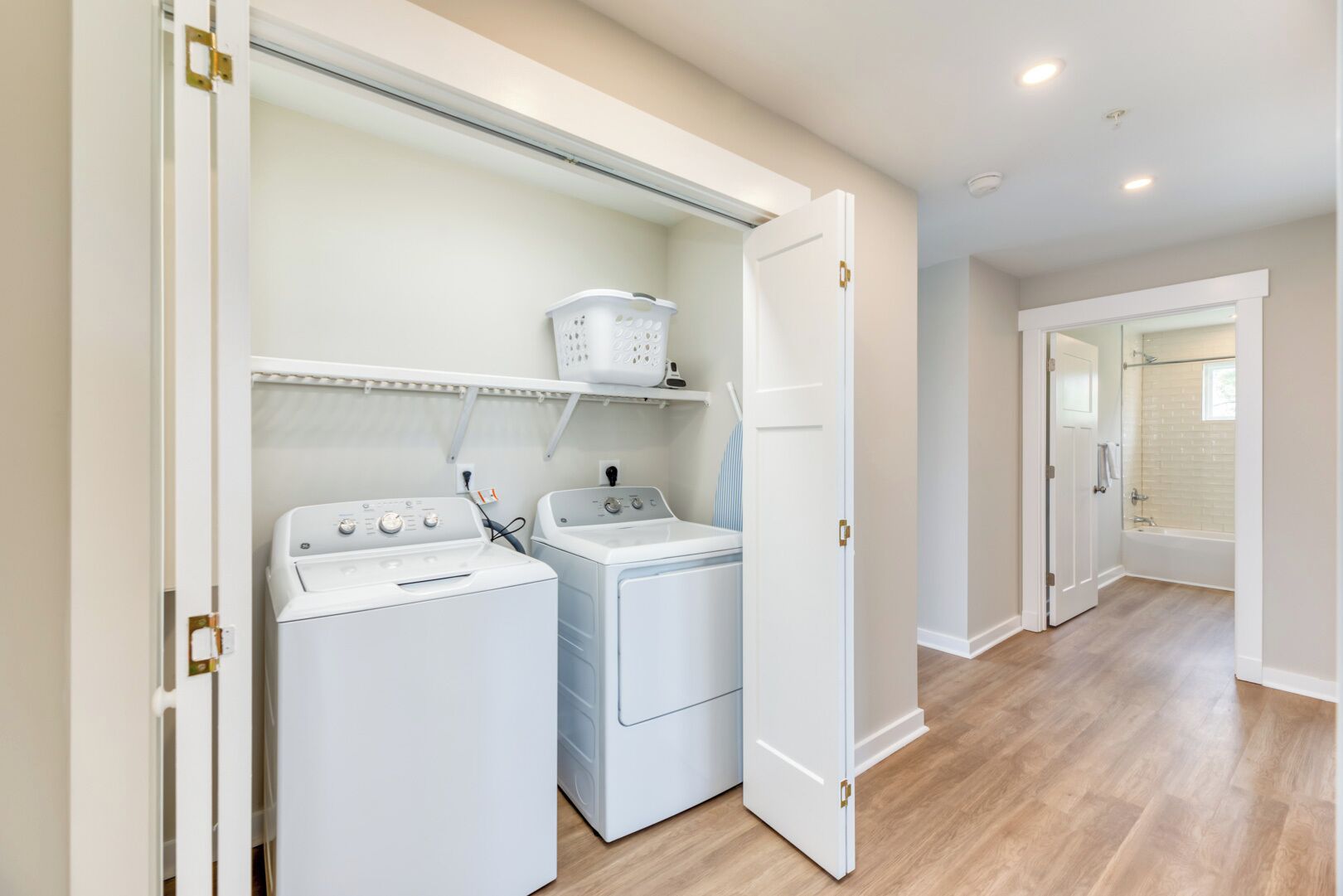 A laundry room with two front-loading washing machines, a drying rack, and a white door leading to a bathroom with a bathtub.
