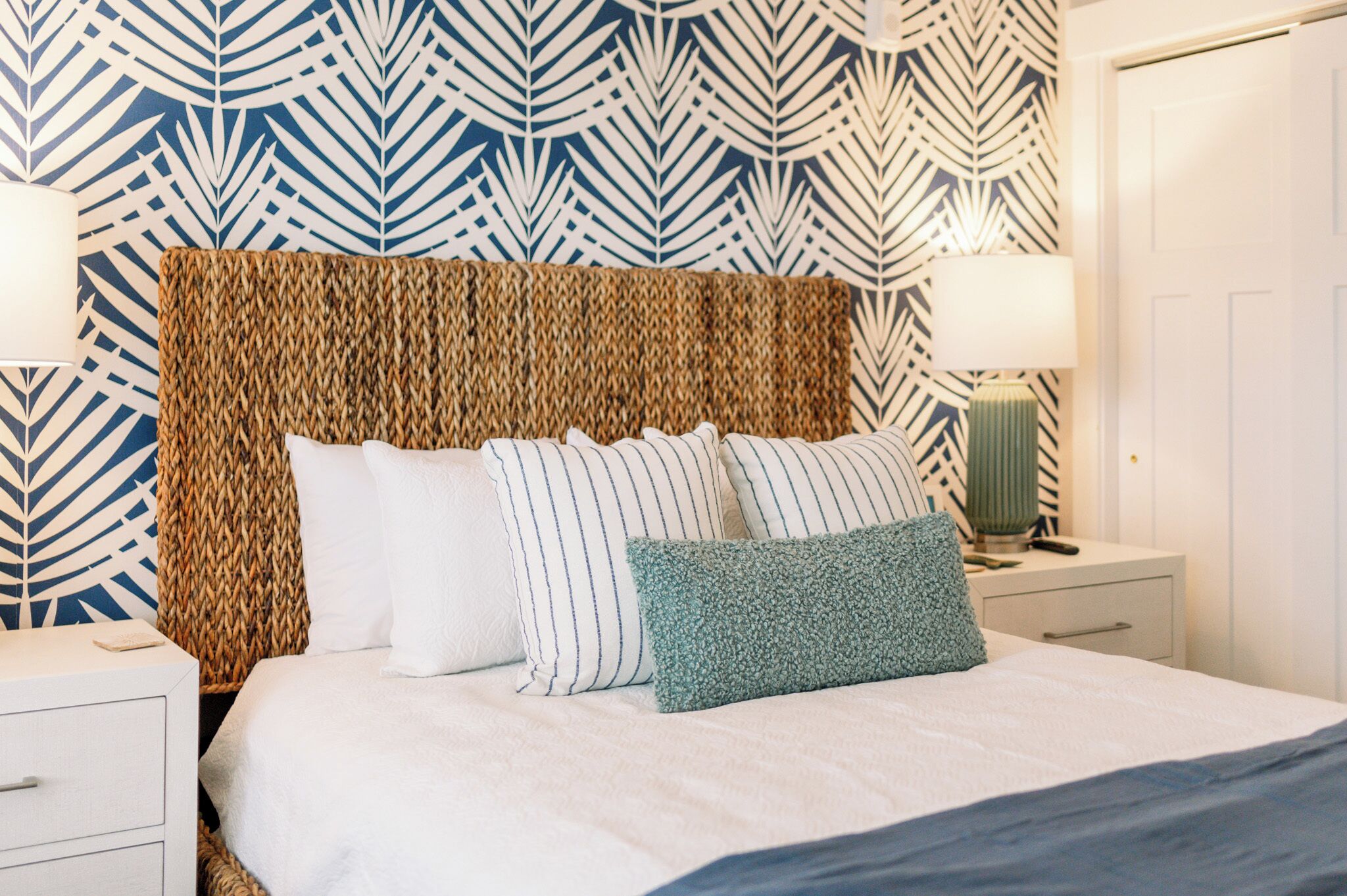Een opgemaakt bed met een geweven hoofdbord, een nachtkastje met een lamp en een behang met patroon.