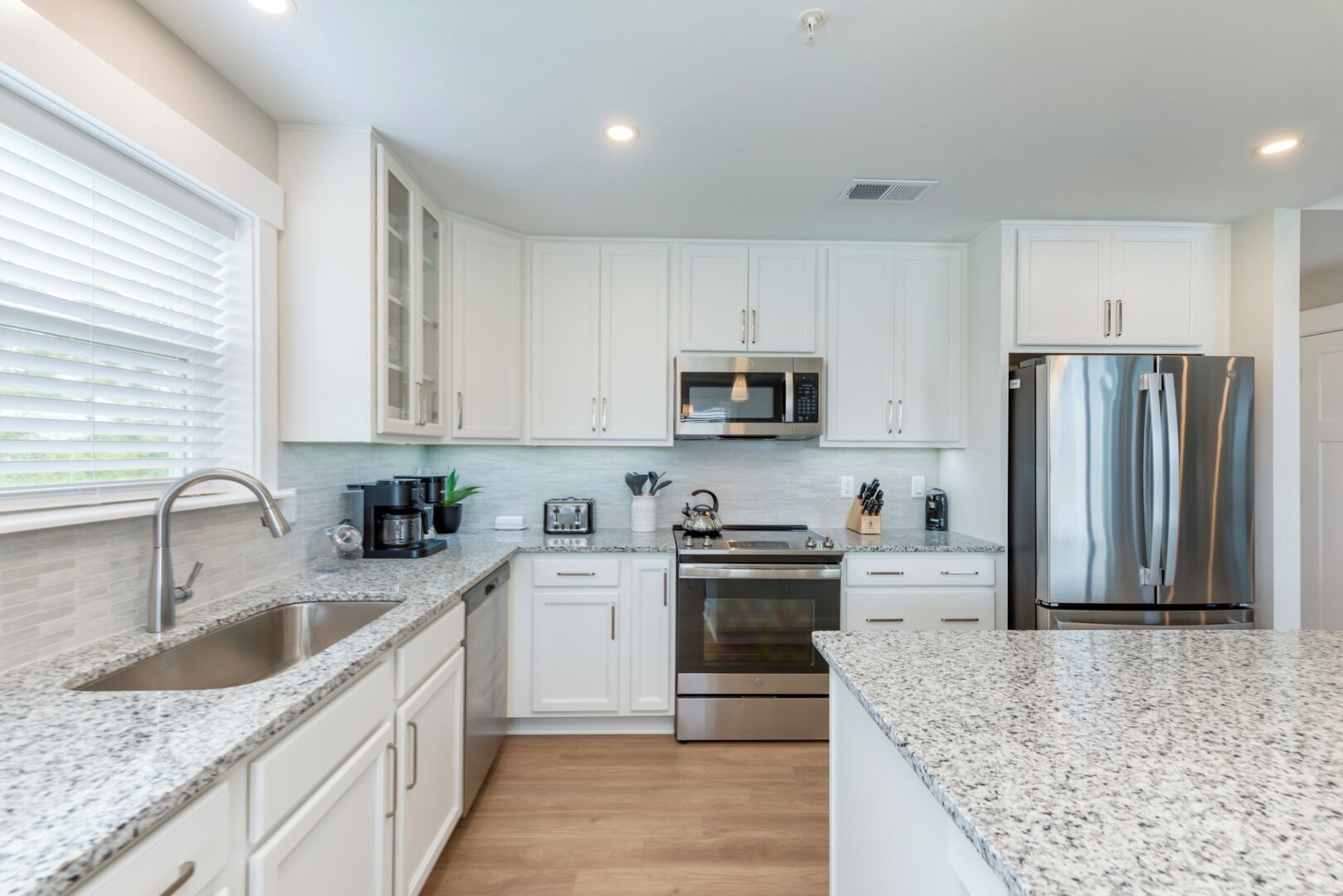A modern kitchen with white cabinets, granite countertops, stainless steel appliances, and a window with blinds.