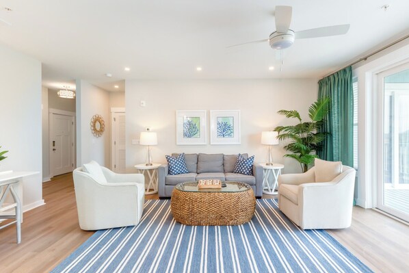 A modern living room with a grey sofa, white armchairs, a woven coffee table, and a ceiling fan. - Hamilton Cay Reserve at Bermuda Bay (Kill Devil Hills)