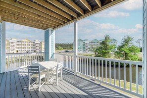 A covered outdoor deck with a table and chairs, overlooking a body of water and a row of buildings. - Hamilton Cay Reserve at Bermuda Bay (Kill Devil Hills)