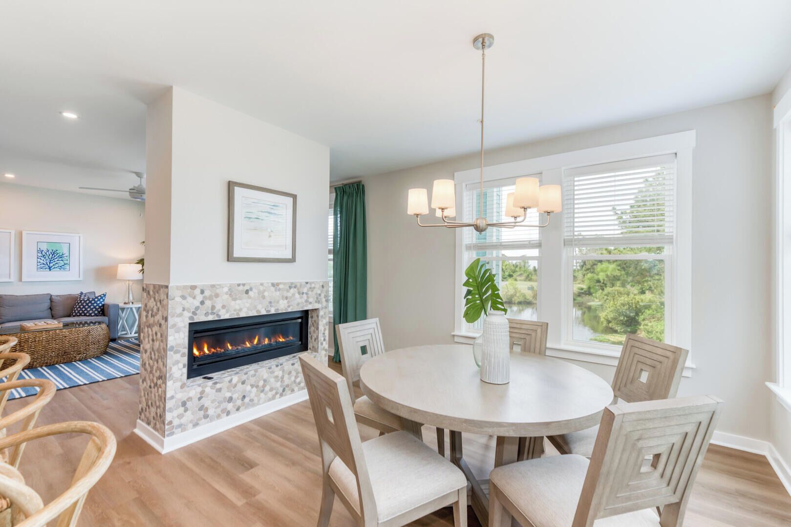 A modern living room with a fireplace, a dining area with a round table and chairs, and a view of greenery through large windows.