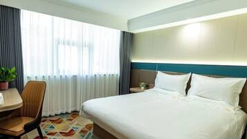 A hotel room with a bed, a desk, a chair, a potted plant, and a window with curtains.