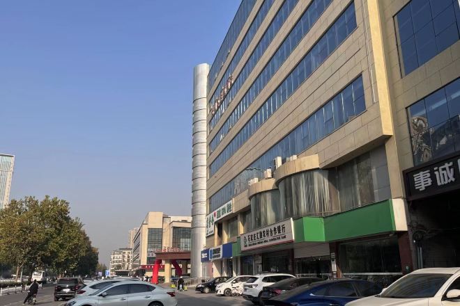 Un immeuble à plusieurs étages, avec des commerces au rez-de-chaussée, des voitures garées le long de la rue et un ciel dégagé.