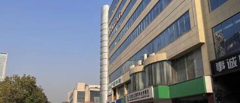 Un immeuble à plusieurs étages, avec des commerces au rez-de-chaussée, des voitures garées le long de la rue et un ciel dégagé.