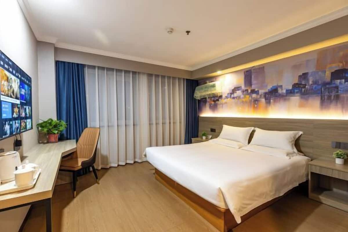 A modern hotel room with a large bed, a desk with a computer, and a cityscape artwork on the wall.