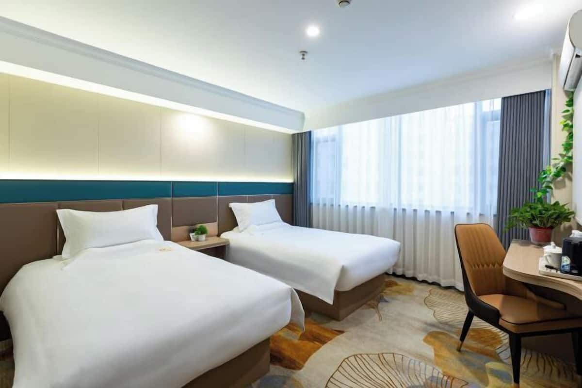 A hotel room with two beds, a desk with a chair, a potted plant, and a large window with curtains.