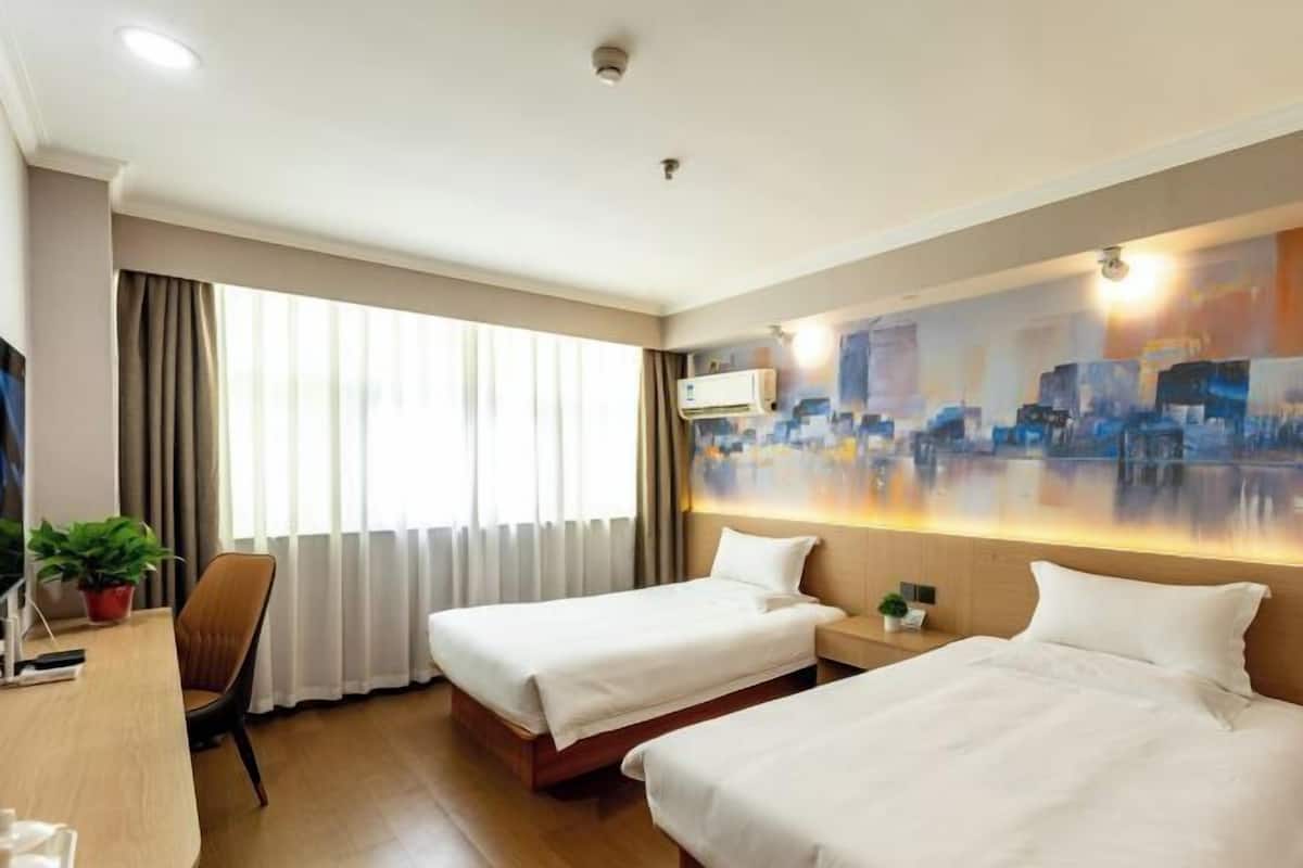 A hotel room with two beds, a desk, a chair, a potted plant, and a cityscape painting on the wall.