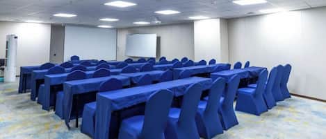 Un salón de conferencias con varias mesas y sillas, una pantalla de proyector y piso alfombrado.