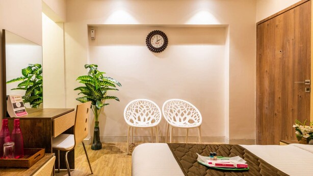 A hotel room with a bed, a desk with a chair, two white chairs, a wooden wardrobe, a wall clock, and a potted plant. - Deluxe Room with Kitchen (Mumbai)