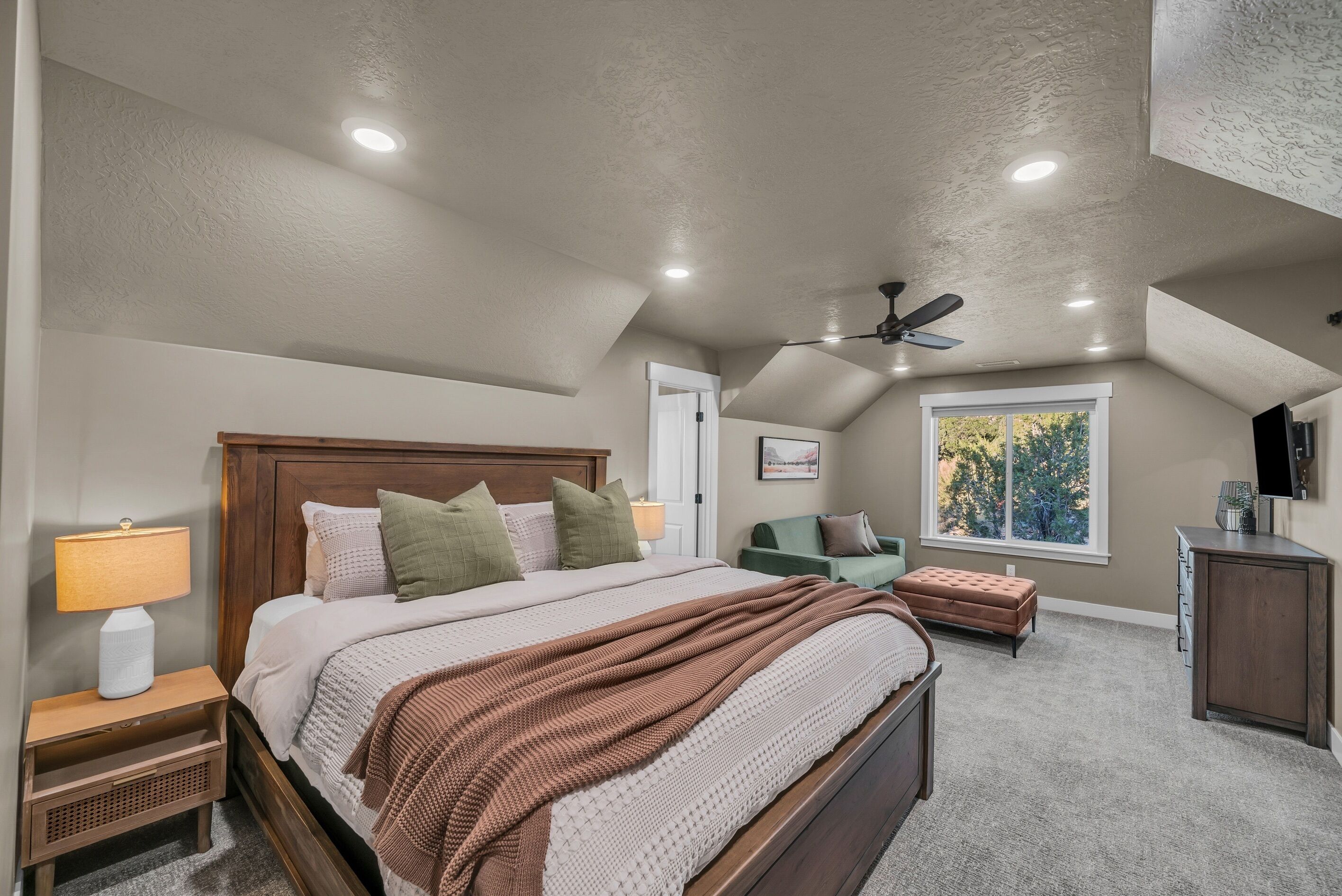 A bedroom with a large bed, a ceiling fan, a window with a view, and a nightstand with a lamp.