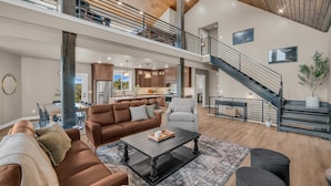A spacious living area with a high ceiling, a staircase, and a kitchen in the background.