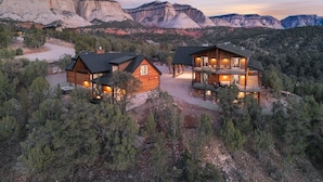 A two-story log cabin with a balcony, surrounded by a forest and mountains.