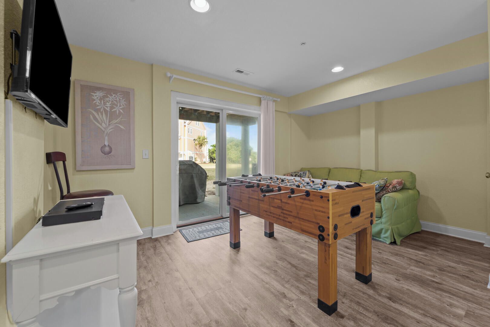 A room with a foosball table, a TV mounted on the wall, a sofa, and a door leading outside.
