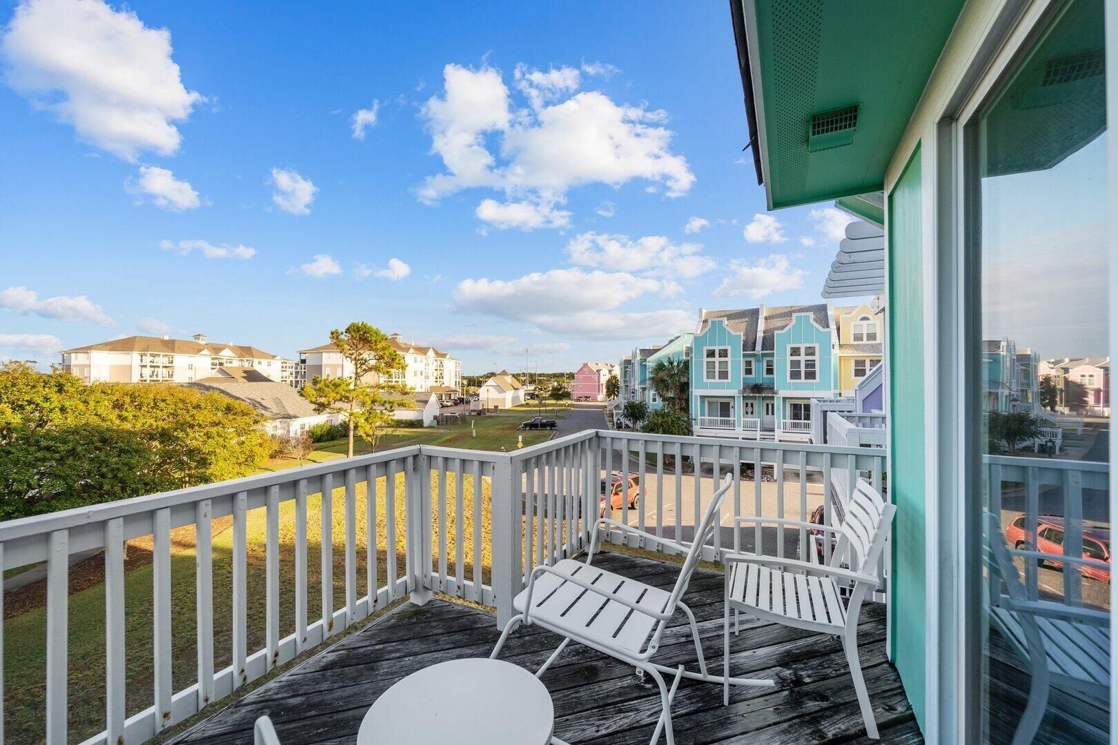 En balkon med hvide metalstole og et lille bord, med udsigt over et boligområde med farverige huse og en klar himmel.
