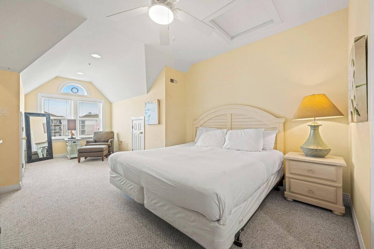 A bedroom with a large bed, a nightstand with a lamp, a chair, and a desk.