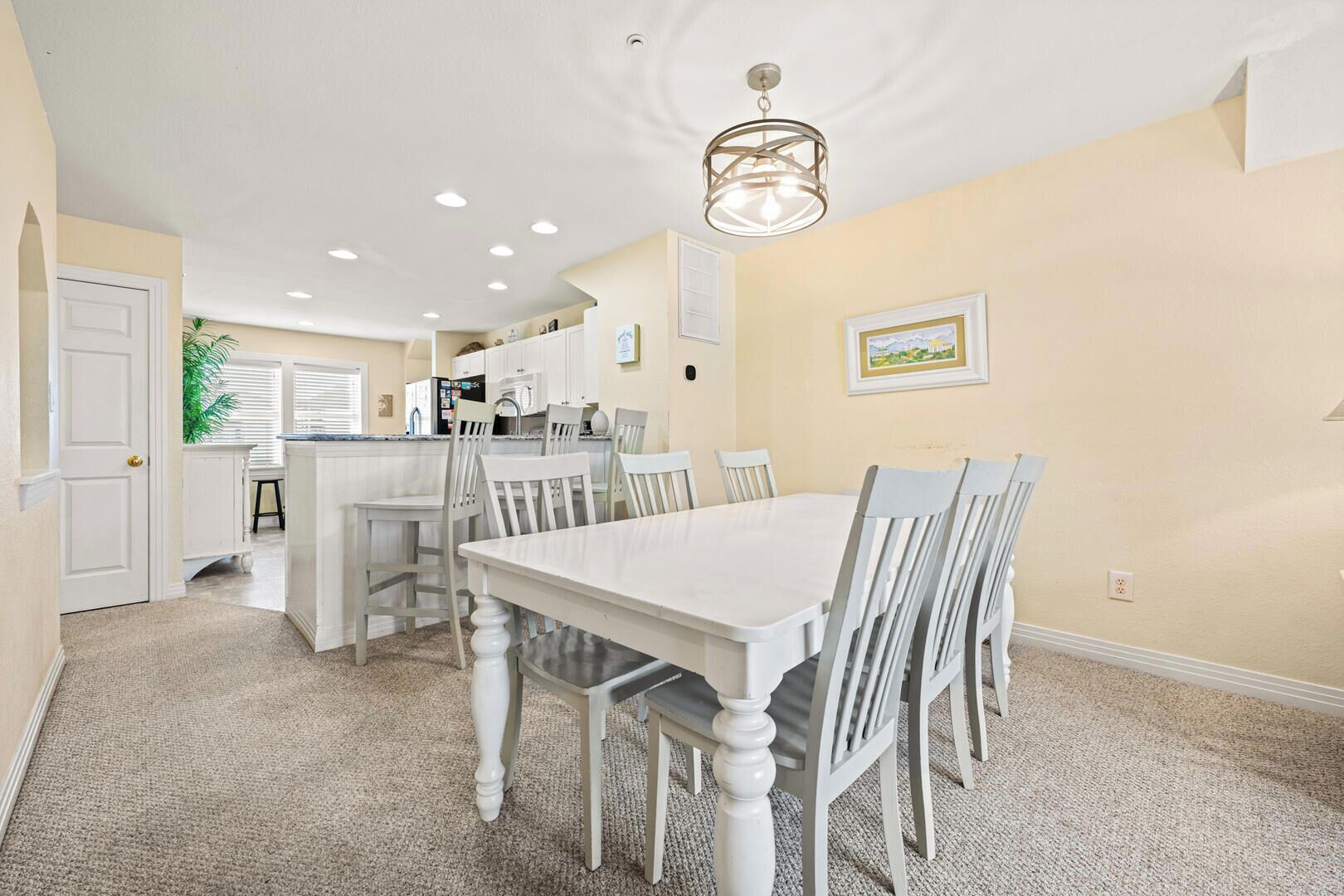 Un coin repas avec une table et des chaises blanches, une cuisine en arrière-plan et un luminaire au plafond.