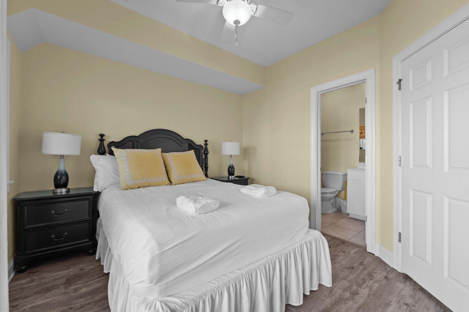A neatly made bed with white linens and two pillows, a bedside table with a lamp, and a ceiling fan.