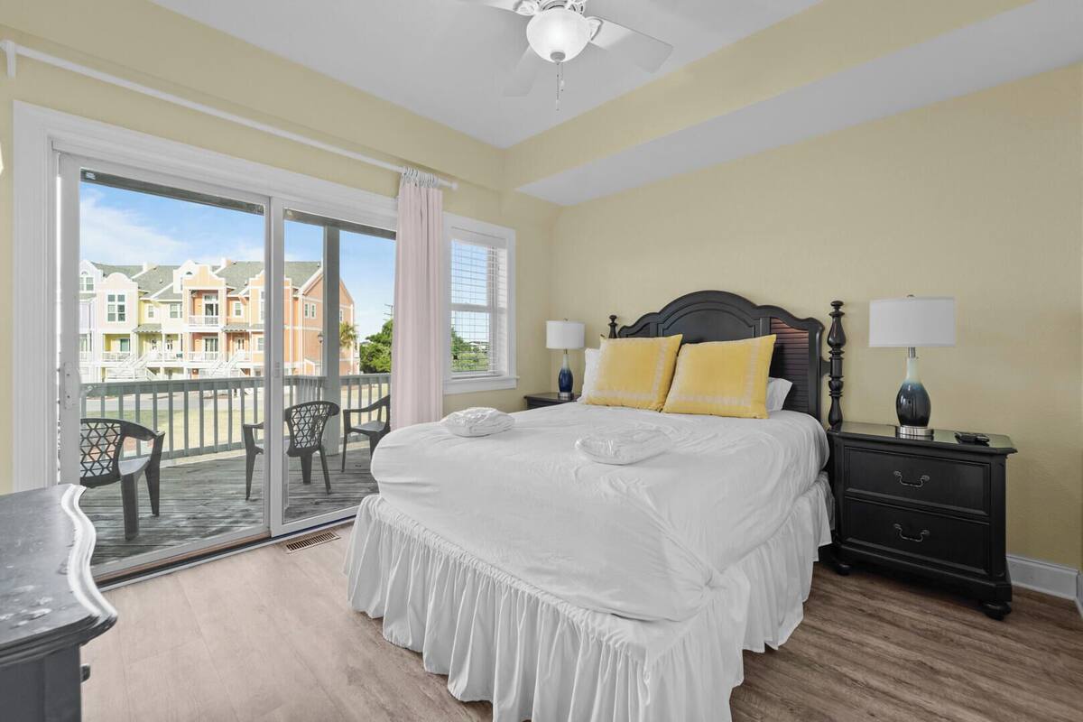 A bedroom with a large bed, a nightstand with lamps, a balcony with chairs, and a view of buildings.
