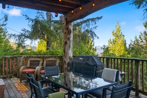 A covered outdoor patio with a glass table and chairs, a grill, and a view of trees and a body of water.