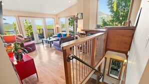 Amplia sala de estar con una escalera de madera, ventanas grandes que ofrecen vistas al exterior y una mesa roja con una planta.