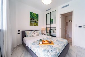 A modern bedroom with a large bed, a painting on the wall, and a tray of fruits on the bed.