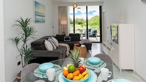 Un salon moderne avec une table à manger en verre pour deux personnes, un canapé, un téléviseur et une vue sur un jardin grâce à de grandes fenêtres.