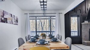 A dining area with a long table set for six, surrounded by chairs, with a view of trees through large windows. - Starlight Chalet | Game Room | Pool Access | AC (Tobyhanna)