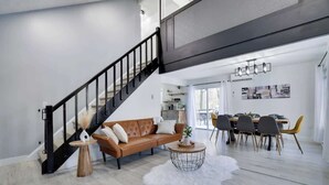 A modern living room with a staircase, a dining area, and a kitchen in the background. - Starlight Chalet | Game Room | Pool Access | AC (Tobyhanna)