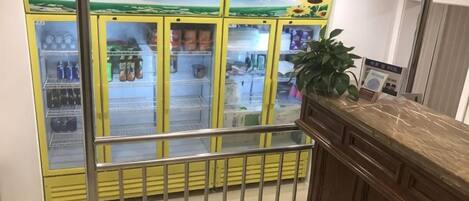 Una máquina expendedora amarilla con varios compartimentos, una barandilla metálica y un mostrador de madera con una planta.