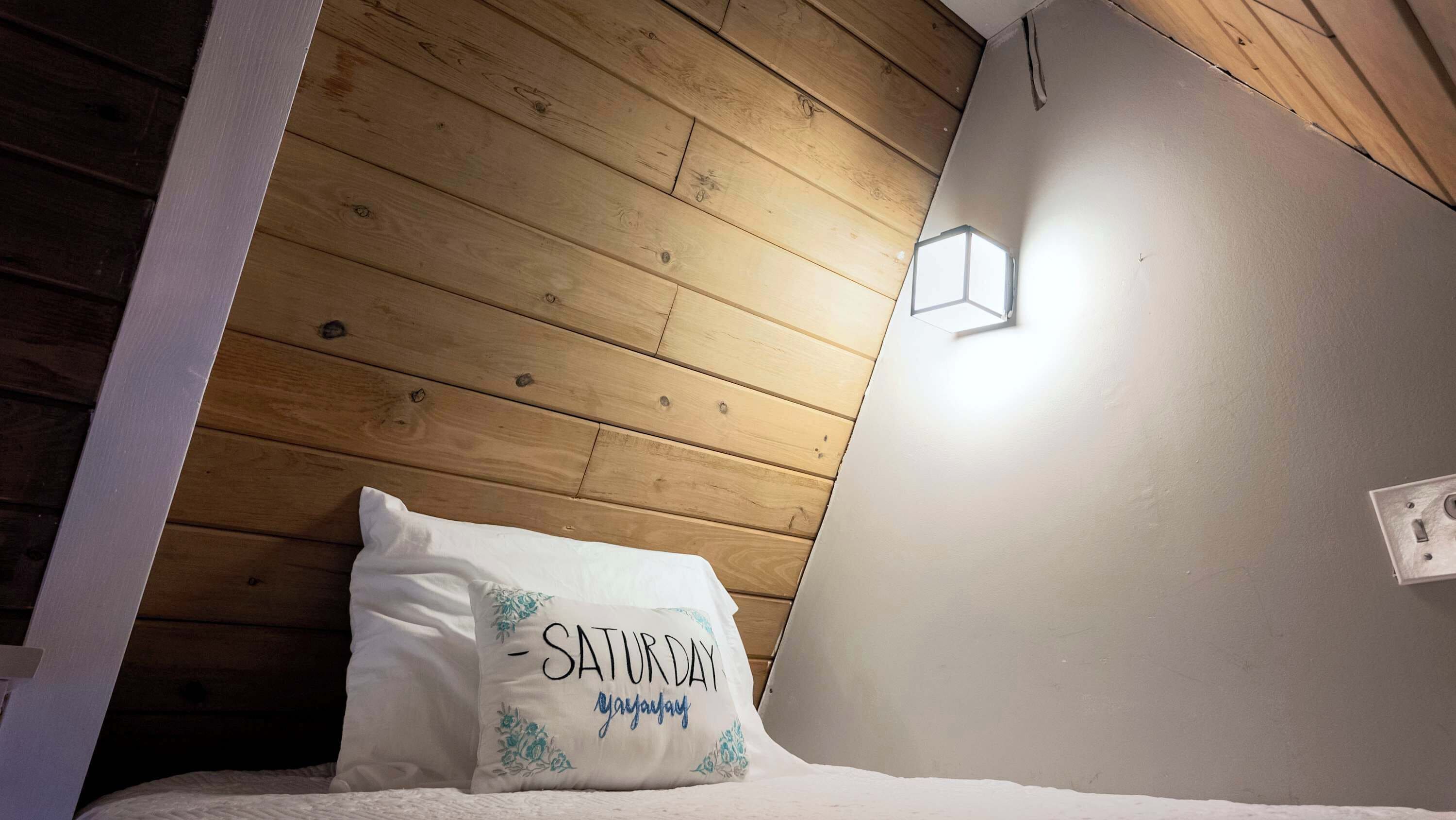 Ein Schlafzimmer mit einem hölzernen Kopfende, einem weißen Kissen mit Schriftzug und einer Wandleuchte.
