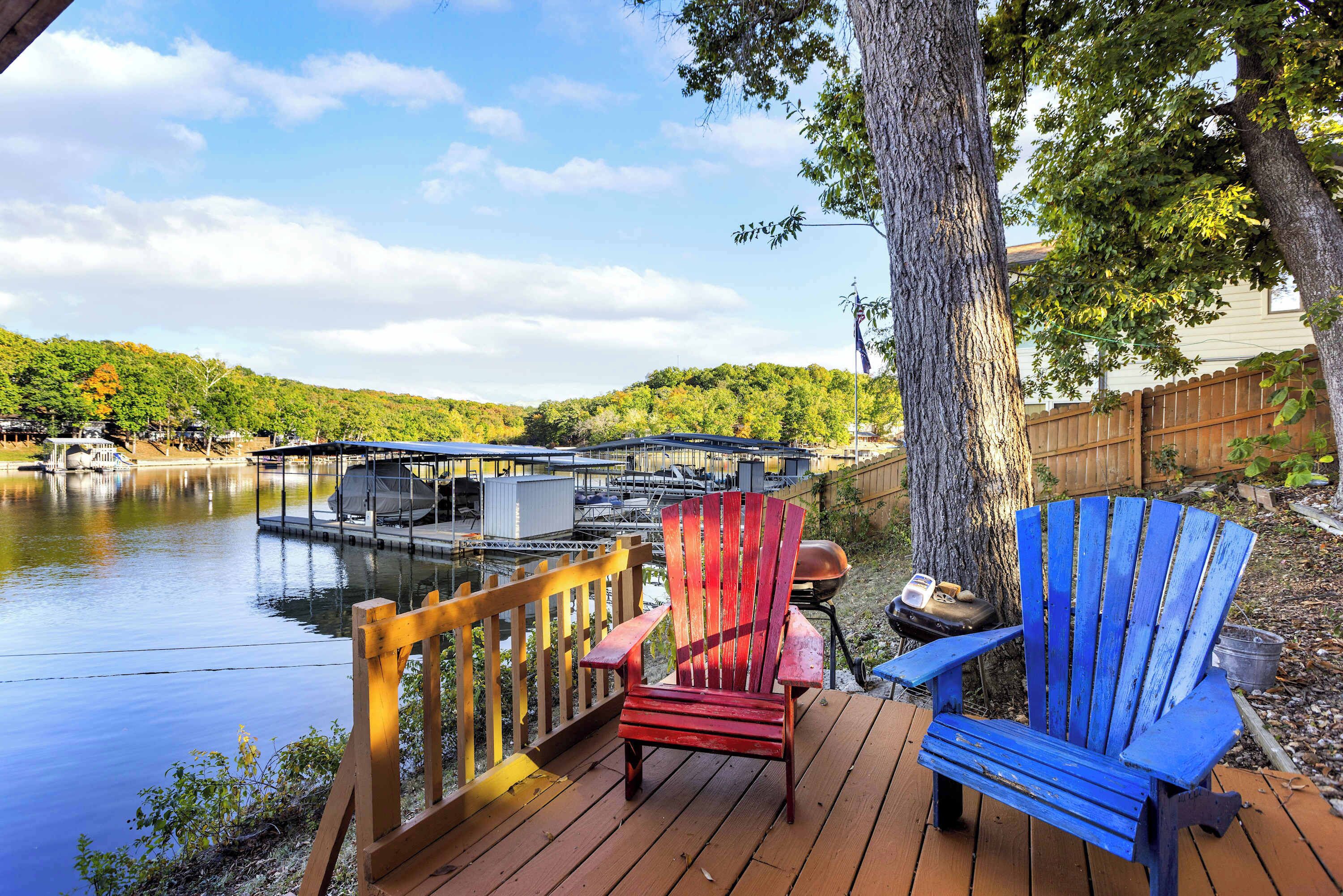 A wooden deck with two Adirondack chairs, a grill, and a view of a lake with boats and docked vessels.