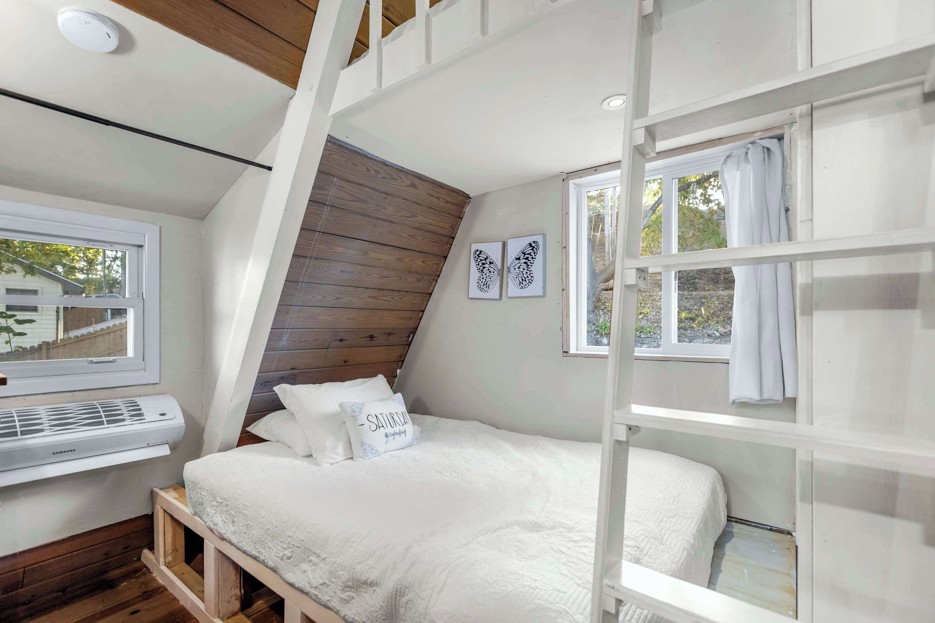 A bedroom with a sloped ceiling, a bed with white bedding, a window with curtains, and a wall-mounted air conditioning unit.