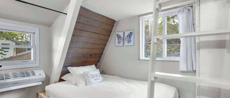 A bedroom with a sloped ceiling, a bed with white bedding, a window with curtains, and a wall-mounted air conditioning unit.