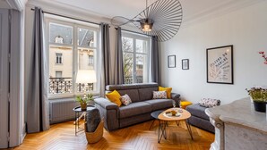 A modern living room with a grey sofa, wooden herringbone flooring, a round coffee table, and a large window with grey curtains.