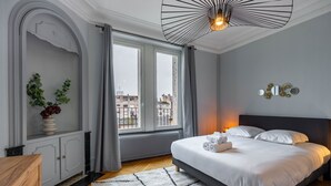 A modern bedroom with a large bed, a view of the city through large windows, a decorative wall piece, and a unique ceiling light fixture.