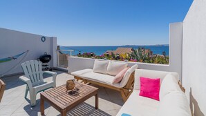 Una terraza con un sofá blanco, una mesa de madera y una silla blanca. Hay una hamaca y una parrilla. La terraza da a un cuerpo de agua y una masa de tierra distante.