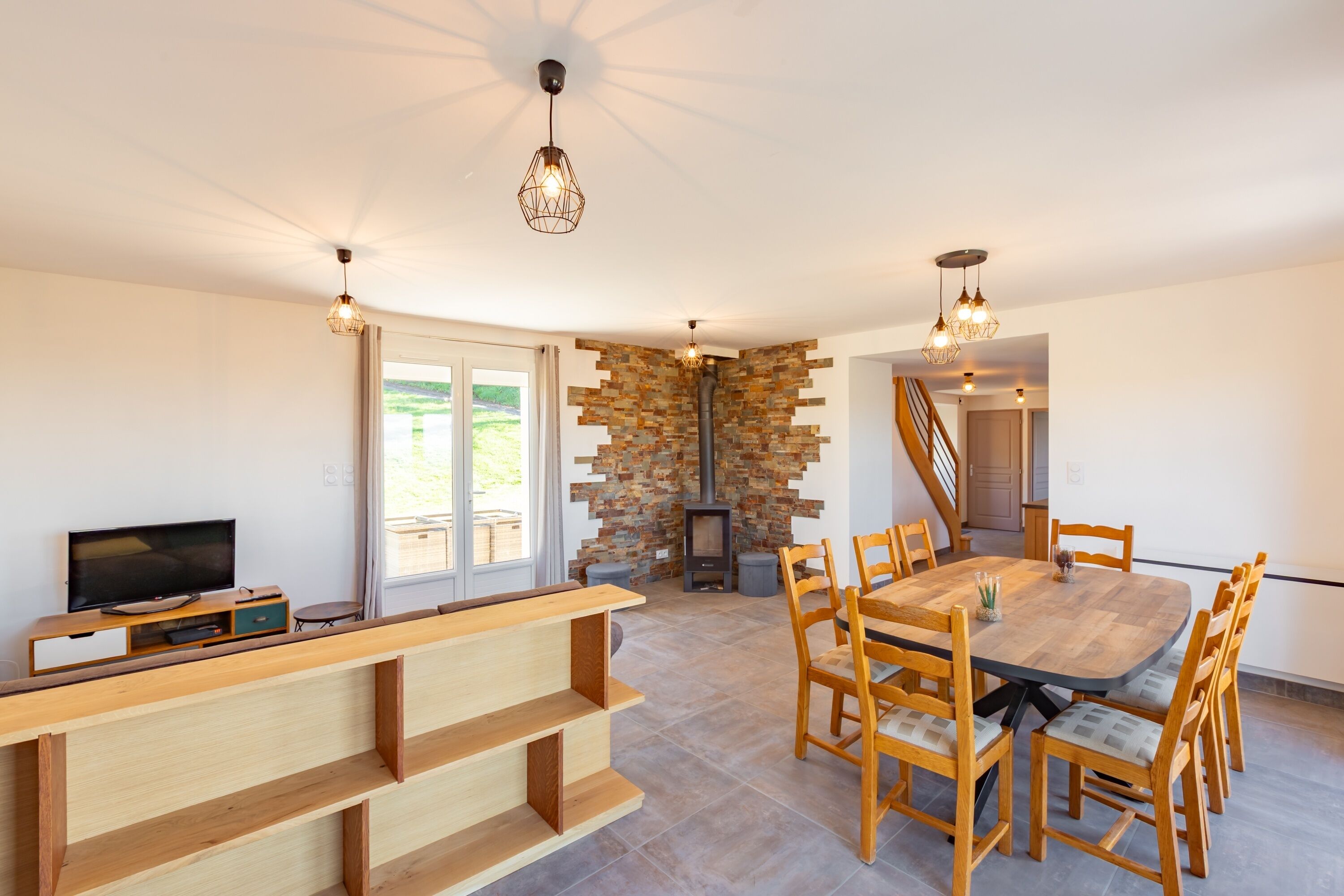 A modern living room with a wooden bench, a TV, a stone fireplace, and a dining area with wooden chairs and a table.