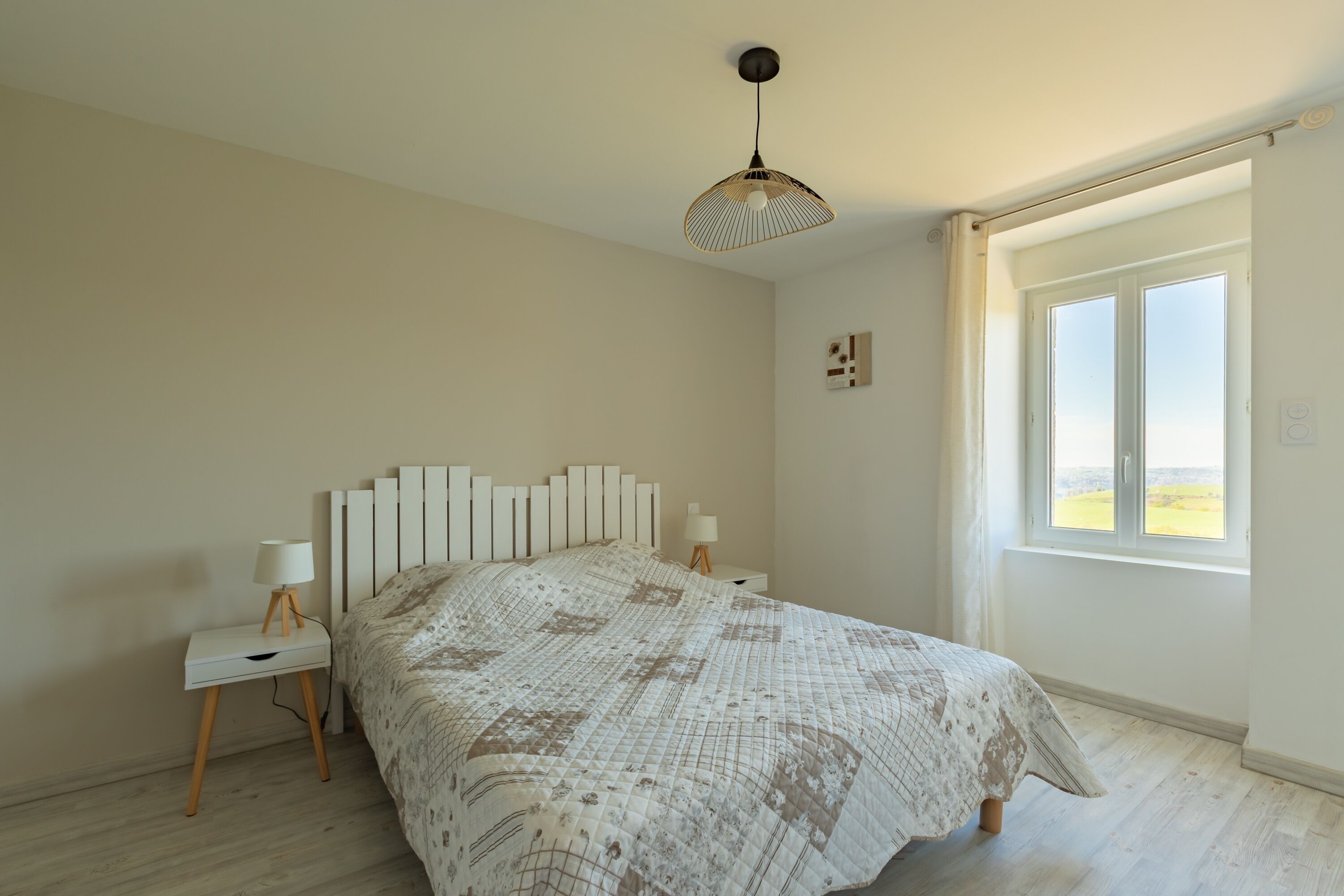 Un dormitorio con una cama grande, dos mesitas de noche con lámparas, una lámpara de techo y una ventana con vistas al campo.