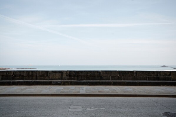 Un sendero pavimentado con el símbolo de silla de ruedas que lleva a una zona costera con vista al mar y a la tierra en la distancia.