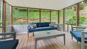 Un área de estar al aire libre techada, con un sofá azul y una mesa, rodeada de piso de madera y ventanas grandes.