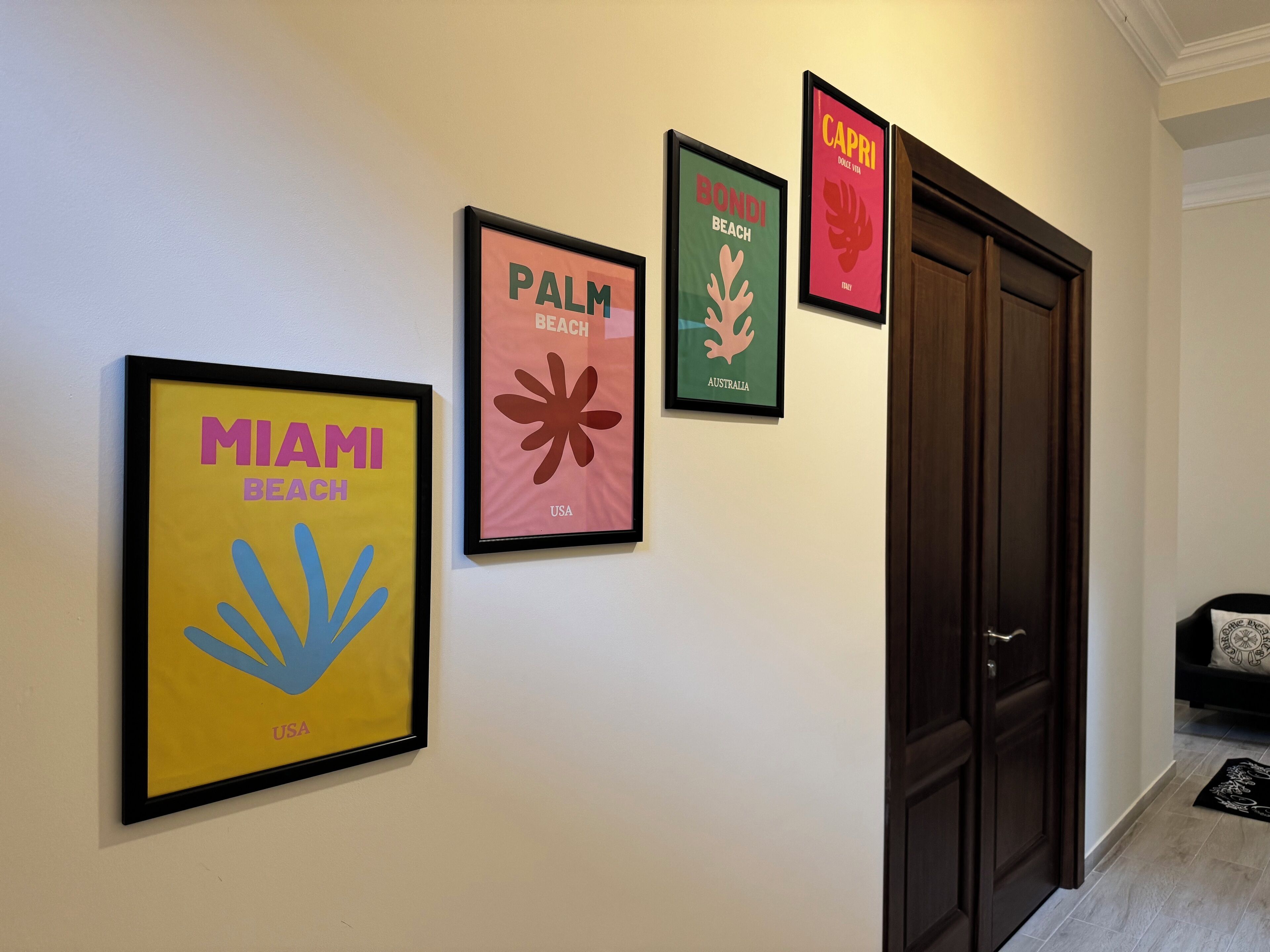 A hallway with four framed posters on the wall, each representing a different beach destination: Miami Beach, Palm Beach, Capri, and Bondi Beach.